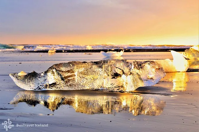 A large block of clear ice glowing gold on wet black sand at sunset