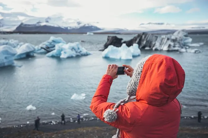 Eine Besucherin in rotem Mantel und Bommelmütze fotografiert blaue Eisberge vom Ufer der Lagune