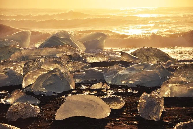 Clear chunks of ice lit gold by a low sun on black sand, with waves breaking behind
